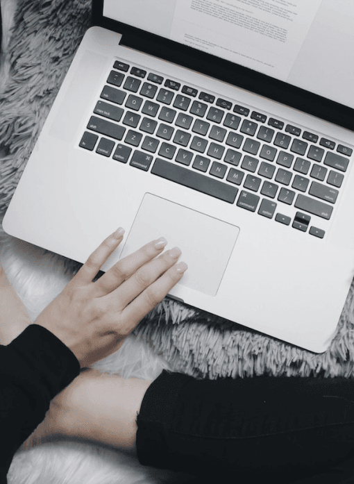Person with laptop on lap, typing on keyboard with document on screen, sitting on gray faux fur rug.