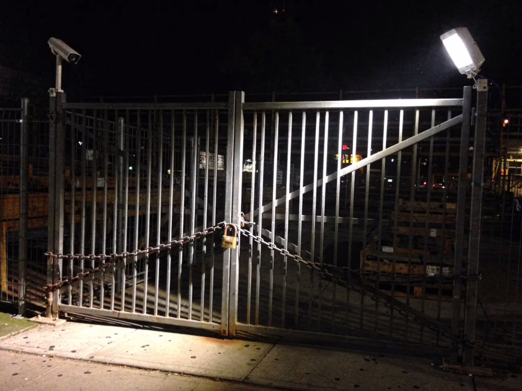 Construction site security San Jose showing a locked perimeter gate with chain and padlock, a security camera on one side and a motion-activated floodlight illuminating the entrance at night