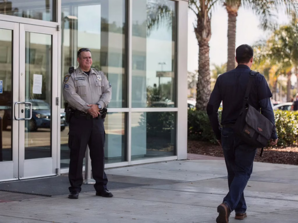 Security guard standing outside glass building as a man walks towards the entrance in daylight.