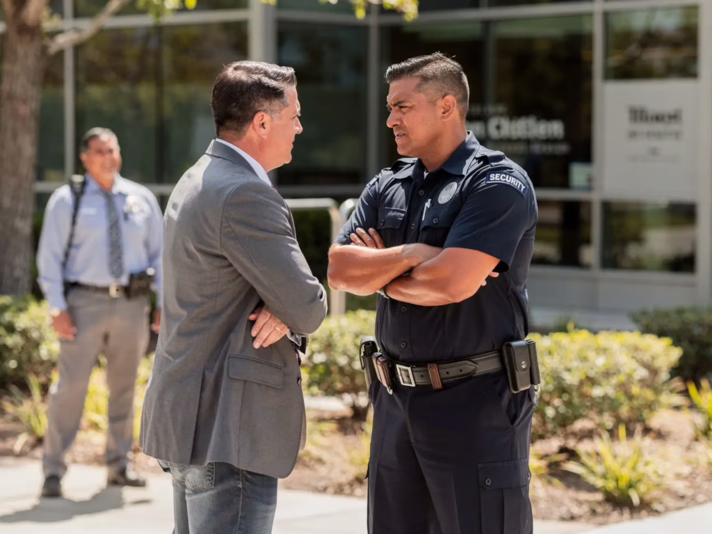 Businessman and security officer talking outside office building on sunny day.